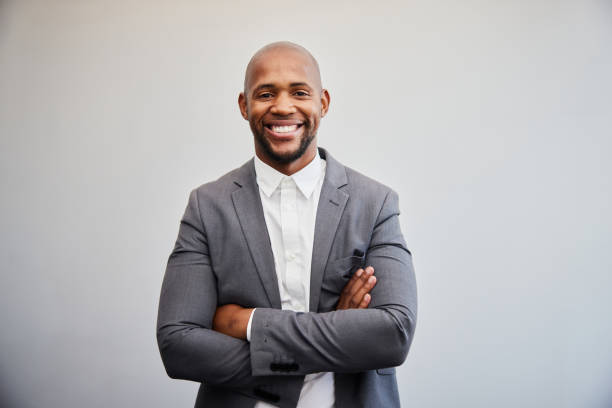 Smiling young businessman standing with his arms crossed against an office wall Portrait of a smiling young businessman wearing a suit standing with his arms crossed in front of a white wall in an office black man stock pictures, royalty-free photos & images