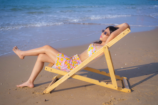 Young Asian woman wearing a swimsuit sits on the beach, sunbathing.