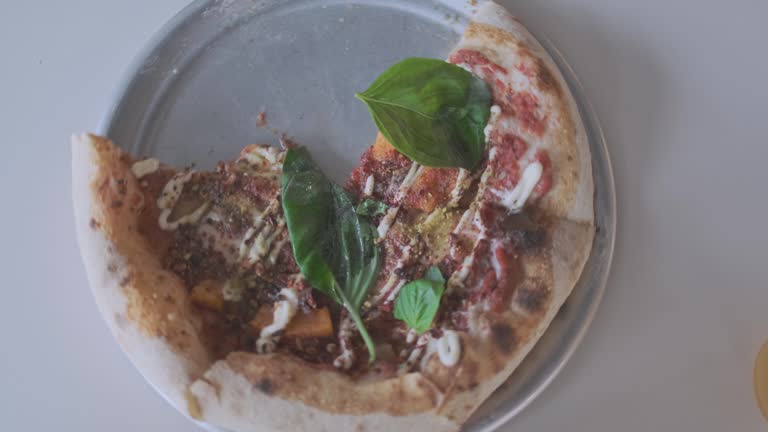 Half-eaten pizza with basil rotates on a plate, top view. A woman's hand rotates a stainless steel plate with a half pizza.