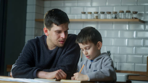 istock Dad help schoolboy with homework at home. Dad helping to son with writing while sitting in the kitchen. Adult parent or tutor with kid boy learning. 2099448850