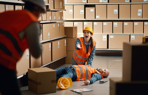 Unconscious injured worker lying on the floor at the warehouse, his shocked colleague is calling for help