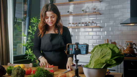 Woman in an apron standing in kitchen records on smartphone new food videoblog. Woman cutting vegetables on a cutting board, blogging, healthy lifestyle, cooking master class event concept