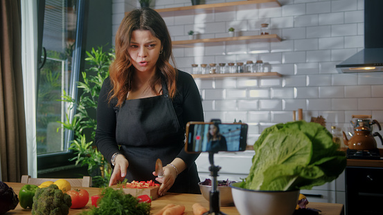 Cute girl in apron standing in kitchen records on smartphone new food videoblog. Woman cutting vegetables on a cutting board, blogging, healthy lifestyle, cooking master class event concept