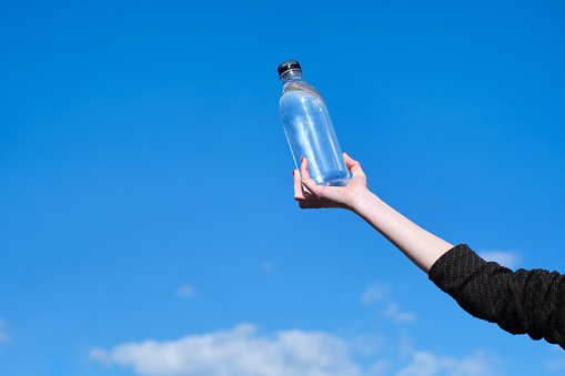 creative, minimalistic, colorful and funny side view of a confident woman hand holding a bottle of water with a blue sky background
