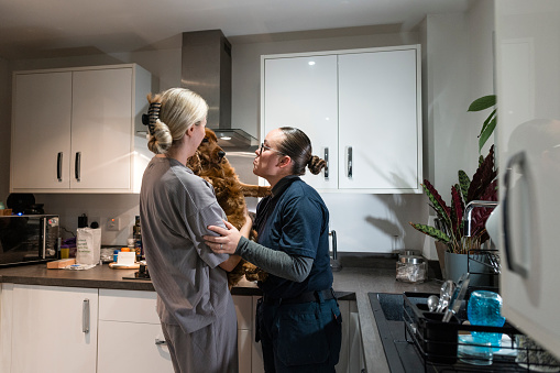 A medium close-up of a same-sex female married couple who are greeting their pet cocker spaniel and giving him kisses. The woman in grey is undergoing IVF Fertility Treatment and is taking hormones intravenously and supplementing the treatment with vitamins.