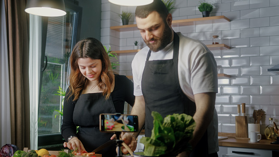 Young couple in an apron standing in kitchen records on smartphone food videoblog. Couple cutting vegetables, preparing healthy a salad. Blogging, healthy lifestyle, cooking master class