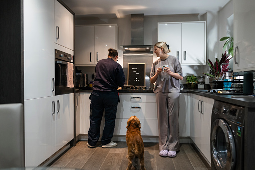 A front view of a same-sex female married couple who are doing their morning routine with their pet cocker spaniel waiting patiently at their feet for his breakfast. The woman in grey is undergoing IVF Fertility Treatment and is taking hormones intravenously and supplementing the treatment with vitamins. They are stood in the kitchen of their family home in the North East of England.