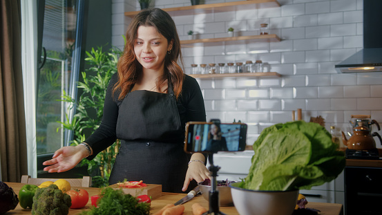 Young woman in an apron standing in kitchen records on smartphone new food videoblog. Woman cutting vegetables on a cutting board, blogging, healthy lifestyle, cooking master class event concept