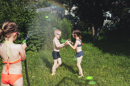 Children boy and girls in a swimsuit plays with splashes of water from a hose on a sunny summer day in the garden. Siblings, friends spend summer holidays, weekends together