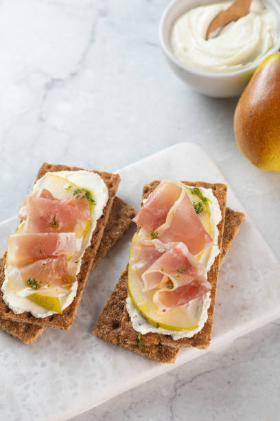 istock Ideal snack wholewheat crispbread with ricotta cheese, prosciutto, sliced pear and thyme on light grey marble cutting board. Healthy snack concept. Italian cuisine 2099573274