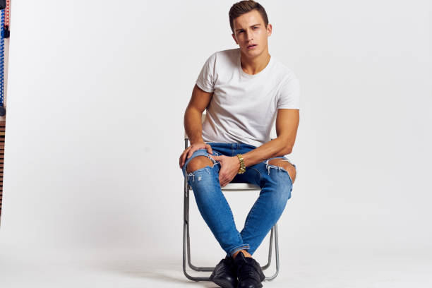 istock Man on a chair indoors torn jeans white t-shirt handsome face model light background 2099598079