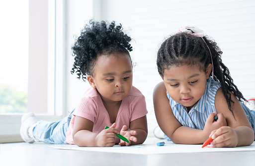African cute little kid girl teaching her sister drawing on paper while lying on the floor. Adorable children have fun drawing with colorful crayons at home. Education, family lifestyles, creativity