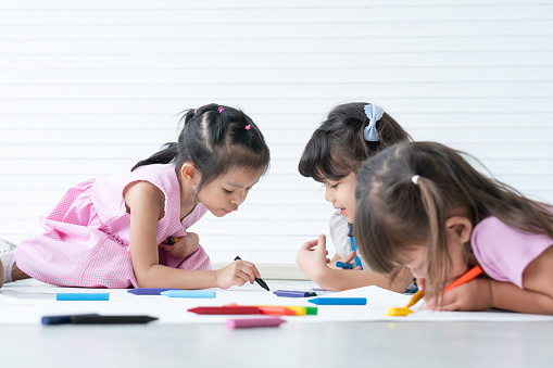 Group of cute little kids girls have fun drawing on paper while sitting and lying on the floor. Adorable happy diversity children drawing with colorful crayons at preschool. Education concept