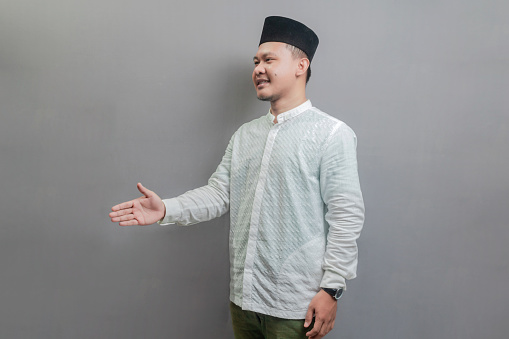 Happy asian muslim man waiving saying hello, friendly welcome gesture wearing a koko shirt and peci with shades of the fasting month, isolated on a gray background