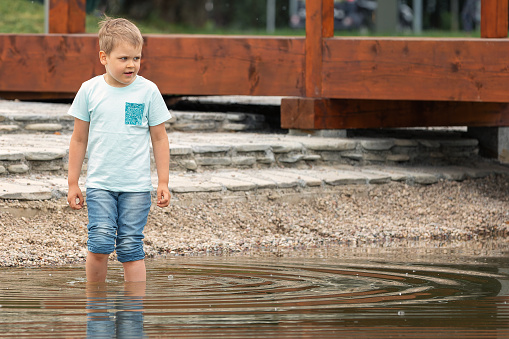 Little boy standing in the water near a wooden linden in a city park.