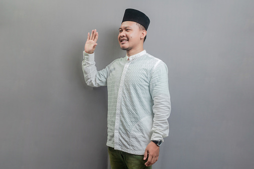 Happy asian muslim man waiving saying hello, friendly welcome gesture wearing a koko shirt and peci with shades of the fasting month, isolated on a gray background