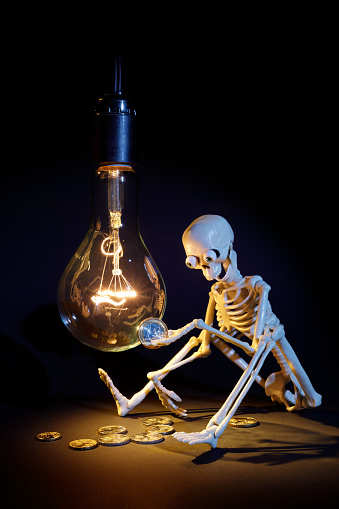 A toy funny human skeleton is sitting in the dark by a hanging big incandescent vintage light bulb, he is holding one euro in his hand and thinking how expensive electricity is. Free space for text.