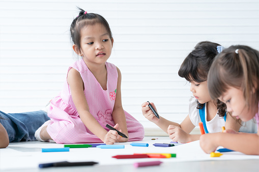 Group of cute little kids girls drawing on paper while sitting and lying on the floor. Adorable happy diversity children drawing with colorful crayons at preschool. Education concept