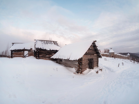 A wooden abandoned house on a snow-covered slope at winter dusk. An old wooden fortress on the slope. Perm Region. Russia.
