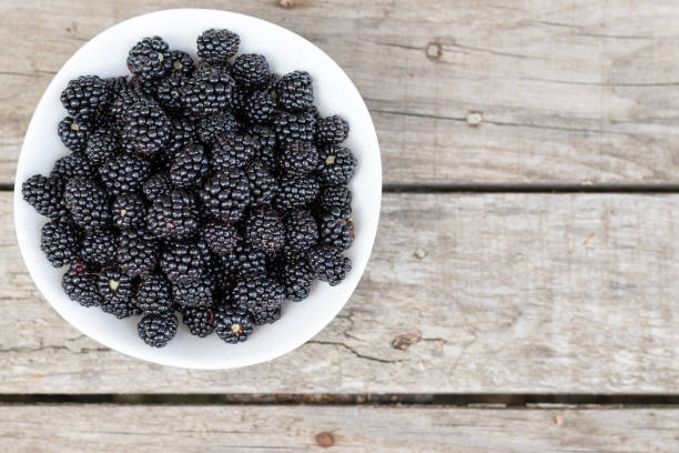 ripe black blackberries in a white plate on a wooden background . View from above. Copy space.