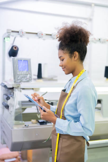 istock Vertical image of a seamstress holding a list of notes and using a pen to check orders for clothing that is being produced by a computerized weaving machine from a clothing manufacturing factory. 2099778406