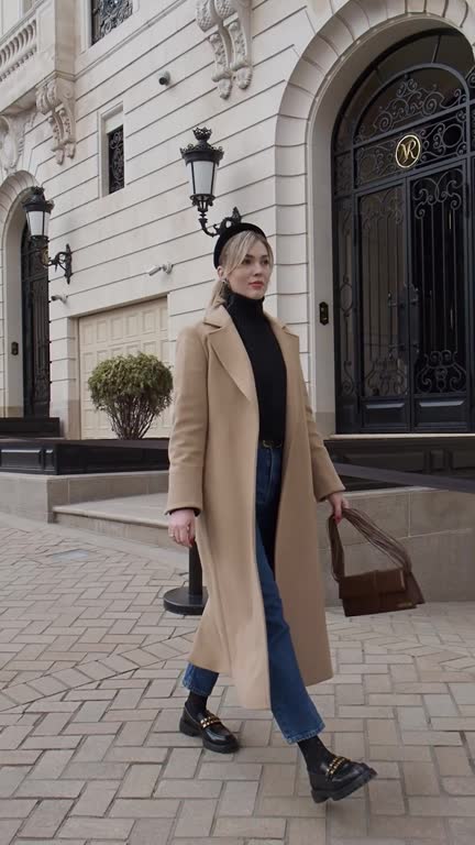 Full length woman walking at historic street of city, against beautiful architecture building, dressed in spring or autumn clothes cashmere beige coat, jeans, turtleneck. Female street style fashion