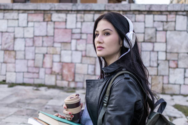 istock Young beautiful female university student with backpack is near  university building. She is wearing headphones and leather jacket while waiting for her college class to start. 2099800645