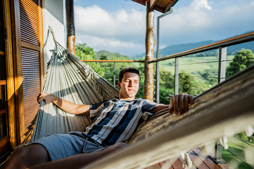 Young man looking away relaxing lying on hammock on balcony farm inn