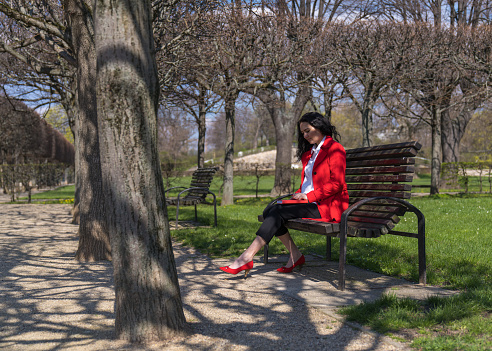 Stylish businesswoman sits on a park bench and writes in a notebook. The lady is wearing a red cloak, white blouse, and red shoes.