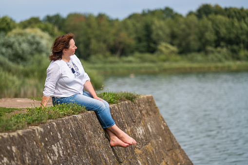 A middle-aged woman enjoys the beautiful landscape. Sits on the edge of the pier overlooking the lake.