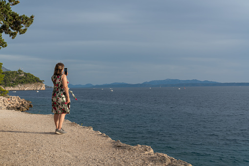 A woman takes pictures of the sea, carabels and a mountain landscape on the horizon on a mobile phone. before sunset