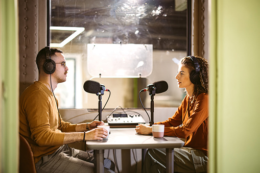 Young coworkers with headphones sitting at table and speaking in mics while recording podcast together in studio
