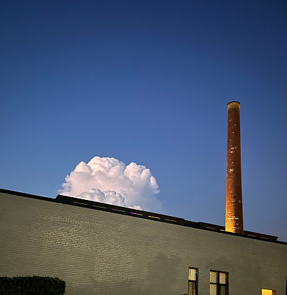 an exterior beige brick wall with an orange smoke stack standing on the top and right of the wall, a billowing rain cloud rising over the horizon against a bright blue sky