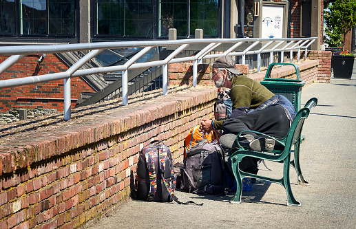 Victoria, Canada – June 09, 2023: A man seated on a bench on a sidewalk staring pensively over a brick wall, his companion is resting on the bench.  In front of him are backpacks.