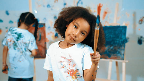 istock Cute girl holding painted brush while student drawing canvas behind. Erudition. 2099970461
