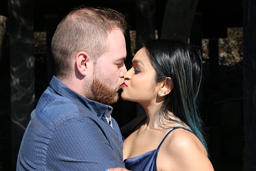 A closeup of a newly engaged mixed race couple. She is wearing long, black, straight hair, makeup, and a blue spaghetti strap dress. He is wearing short brown hair and beard and a blue shirt.