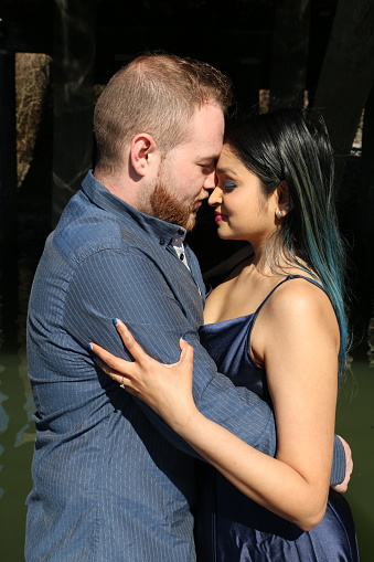 A newly engaged mixed race couple embracing each other. She is wearing long, multi colored, straight hair, makeup, jewelry, a blue spaghetti strap dress. He is wearing short brown hair and beard, and a blue shirt.