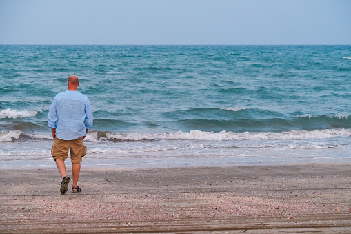Back view of an big man tourist,  walking on sand beach towards waves on Arabic sea, Oman.