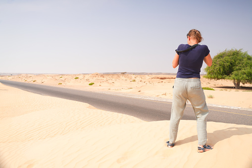 Back view of an senior woman, photographer, waiting for photo moment on top of small sand dune by desert road in south of Oman.