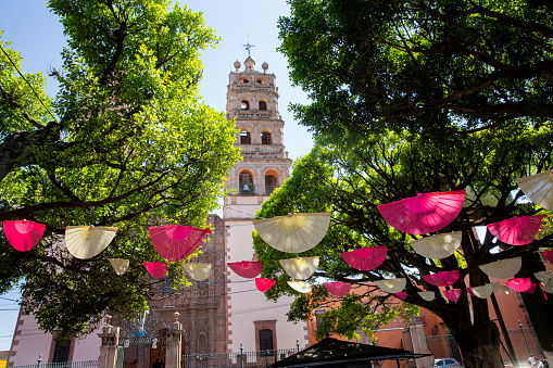 One of the bell towers of the Nuestra Señora de la Luz parish church, seen across the central garden and decorative paper flags in the historic center of the town of Salvatierra in the state of Guanajuato