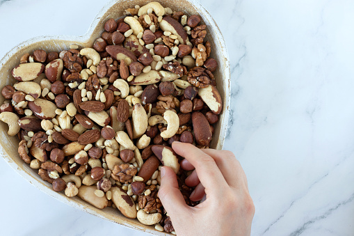 Hand holding Brazil nut and heart-shaped bowl full of mixed nuts isolated on white marble background. Top table view. Close-up. Healthy snacking, eating organic food concept.
