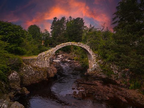 Vibrantly coloured clouds above old stone remains of an arched Coffin Bridge. Lush greenery surrounding rustic arch of an ancient Carrbridge Packhorse Bridge that crosses a serene mountain stream.