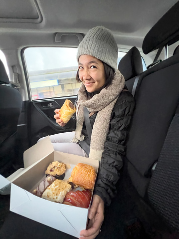 Sustainable eating- a multiracial young woman holds a vegan sausage roll and a compostable cardboard box of vegan pastries including croissant, chocolate and carrot cake, rolls, and muffin from a local specialty bakery.  Vancouver, British Columbia, Canada.