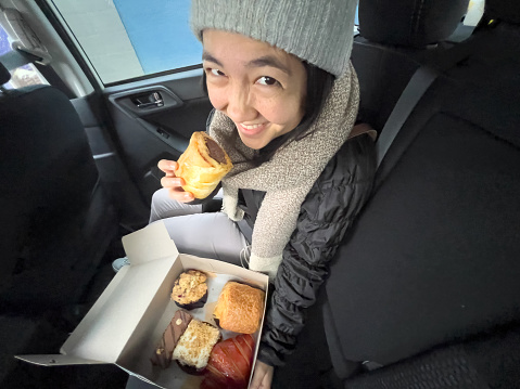 Sustainable eating- a multiracial young woman holds a vegan sausage roll and a compostable cardboard box of vegan pastries including croissant, chocolate and carrot cake, rolls, and muffin from a local specialty bakery.  Vancouver, British Columbia, Canada.