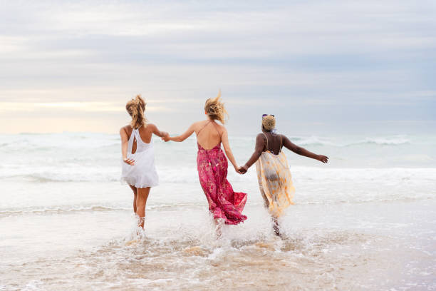 group of three female friends of different races (Latina, white and black) enjoy a day at the beach running together and holding hands along the seashore on a beach.