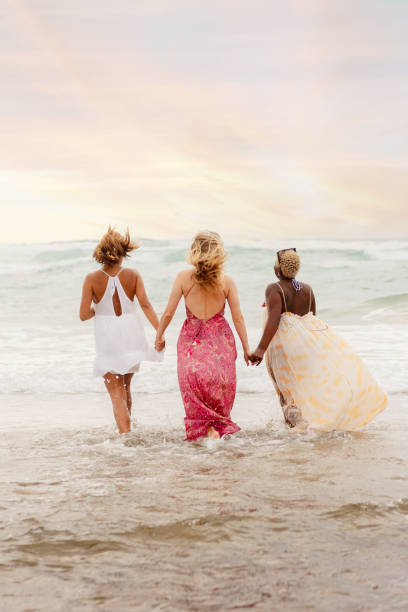 Vertical photograph of a multiracial group of three friends holding hands cooling off on the seashore during a summer day