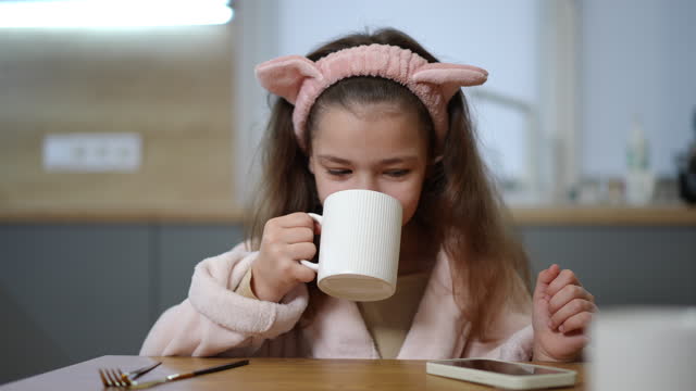 Slow motion. A girl, in a bathrobe and with a hairband on her head, sits at a table in the kitchen and drinks from a mug in her hand. The girl smiles while looking at the screen of her mobile phone.