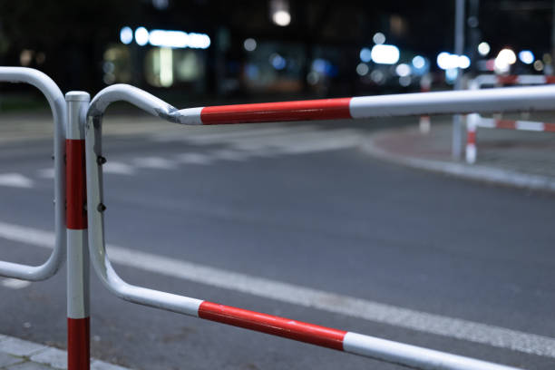 This image depicts a road barrier situated on a street in Arrecife. The barrier, made of orange plastic with vertical slats, serves as a physical obstruction to traffic. Affixed to the left side of the structure is a red circular “No Entry” sign featuring a white horizontal stripe, clearly indicating restricted access. The asphalt road, marked with yellow lines, suggests urban infrastructure, while the background includes a sidewalk with stone tiles and a textured wall. The image conveys a sense of controlled traffic management, reflecting standard measures used to regulate movement and ensure public safety in city environments.