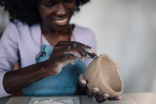 Cropped picture of a multicultural pottery course student sitting in studio and decorating handmade earthenware. Close up of african american woman's hands decorating creative handcraft and pottery.