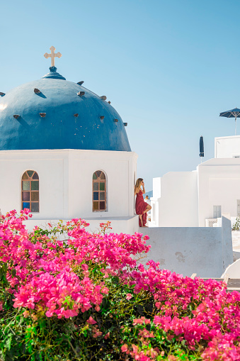 Young beautiful stylish happy female model tourist lady walking and enjoying the amazing view in a red color long red dress looking at the view of the famous traditional and iconic typical traditional blue dome church and colorful bougainvillea flowers at the beautiful sunset in Greek Santorini Island, Greece Oia village, Greece